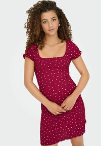 Jeune femme avec des cheveux bouclés portant une robe ajustée à manches courtes de couleur bordeaux avec des pois blancs, se tenant devant un fond uni.