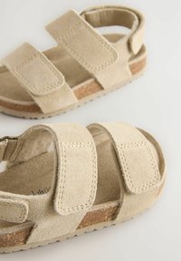 Beige Wildledersandalen mit zwei verstellbaren Riemen, genähten Akzenten und einer strukturierten Korksohle. Nahaufnahme, die das Material und das Design hervorhebt.