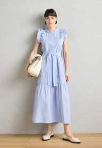 Maxikleid in Blau- und Weißstreifen mit Rüschenshirts, Knopfleiste und Taillengurt. Kombiniert mit einer beigen Handtasche und flachen Schuhen.