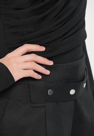 Dłoń oparta na czarnych spodni high-waisted z szerokim paskiem z trzema srebrnymi zatrzaskami, w połączeniu z marszczoną czarną bluzką.