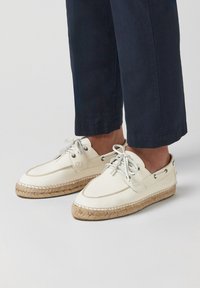 Zapatos de barco de cuero blanco con suela de esparto texturizada, cordones grises y ojales plateados. Presenta detalles de costura a lo largo de los bordes.