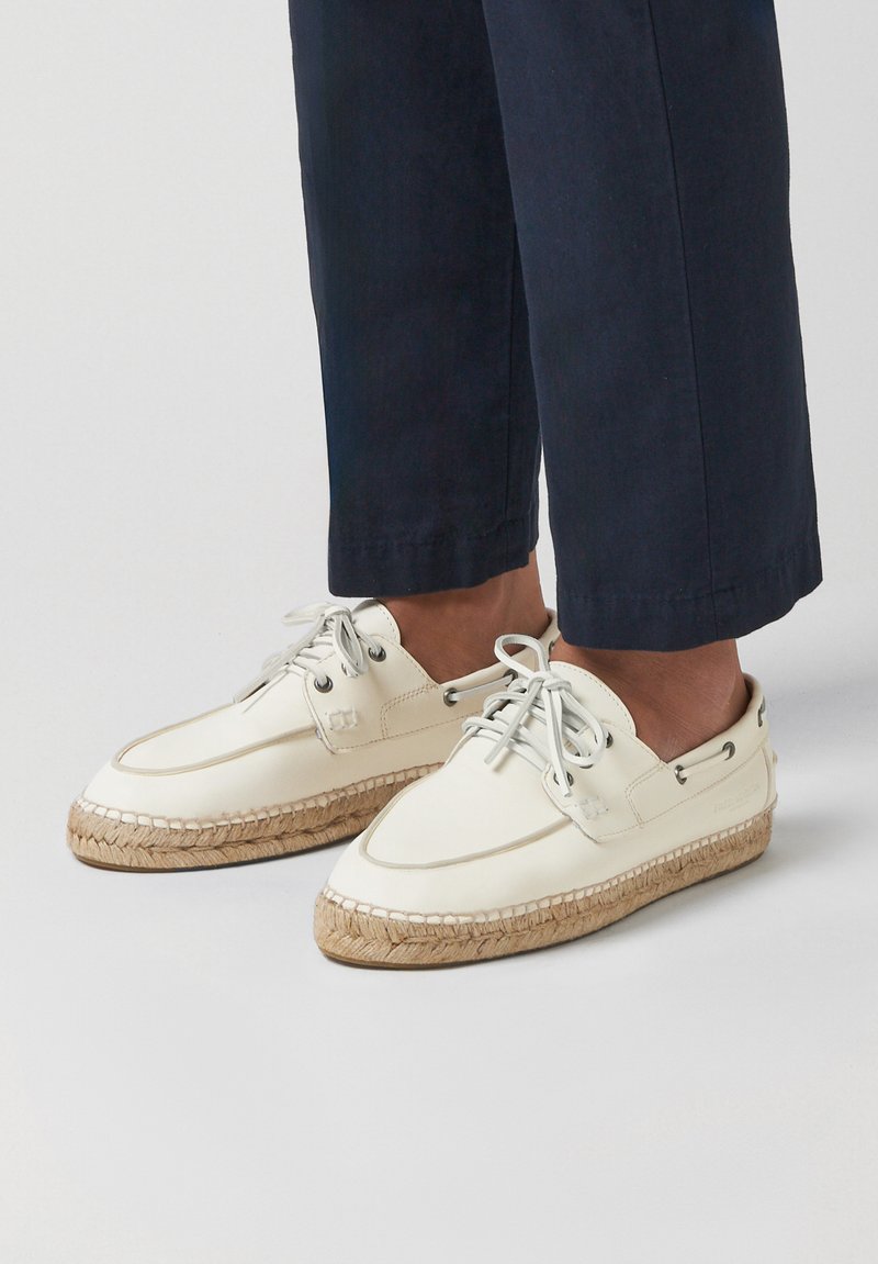Weiße Espadrille-Schuhe aus Leder mit gewebten Jutesohlen. Sie verfügen über runde Nähte, Metallösen und Schnürsenkel. Getragen zu marineblauen, gekürzten Hosen.