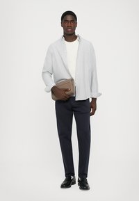 Young man wearing navy pants, white shirt, light gray striped button-up shirt, black shoes, holding a beige clutch bag against a white background.