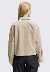 Beige cotton jacket with a collared neck, dropped shoulders, and buttoned cuffs, worn over dark denim. Smooth texture and cropped fit.