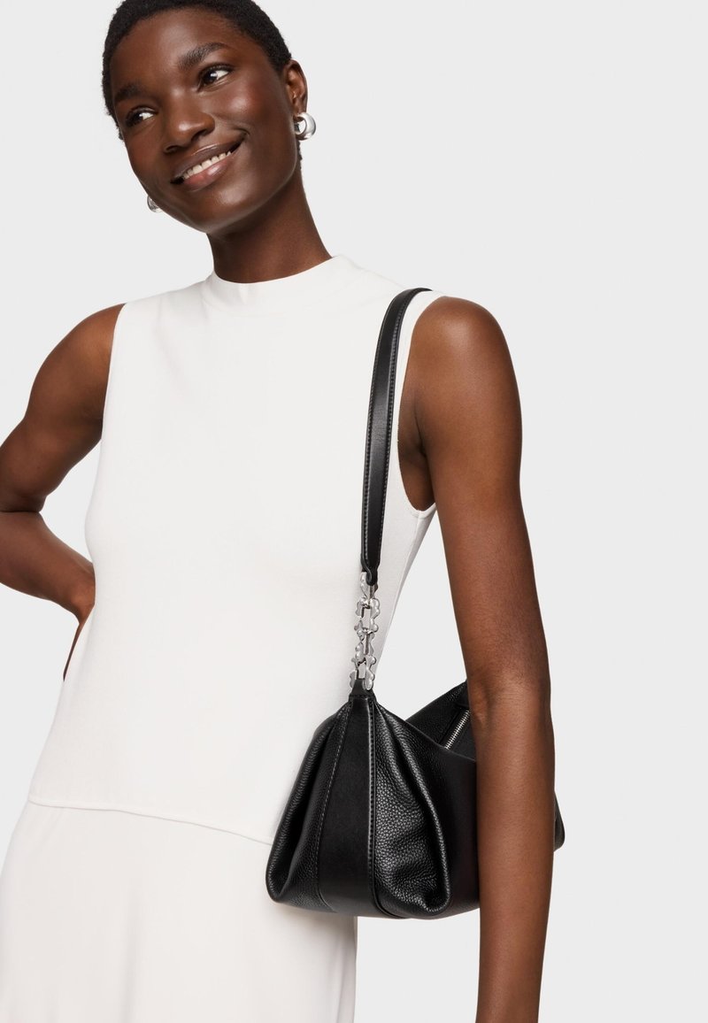 Czarna torebka ze skóry o teksturowanej powierzchni i luźnym fasonie, z srebrnym łańcuszkowym paskiem, trzymana na tle białej, bezrękawnikowej bluzki.