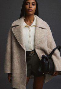 Žena v béžovém volném vlněném kabátu, bílém zapínacím svetru, černých šortkách a držící černou koženou kabelku na tmavém pozadí.