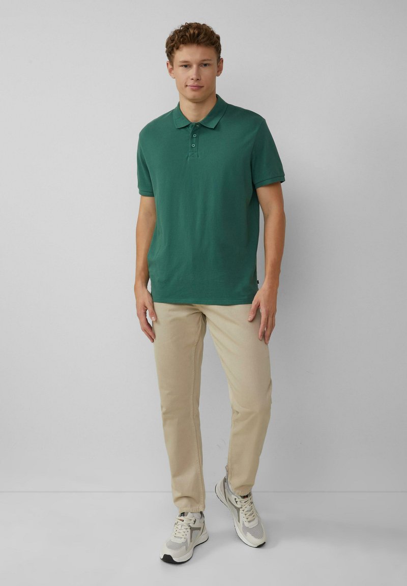 Polo en coton vert avec col et patte de boutonnage à trois boutons, associé à un pantalon beige et des baskets grises. Design simple avec des manches courtes.