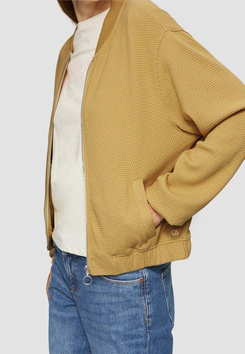 Personne portant une veste zippée jaune moutarde texturée par-dessus un t-shirt blanc et un jean bleu, avec une main dans la poche de la veste.