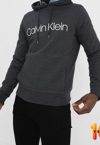 Felpa con cappuccio grigio scuro con logo "Calvin Klein" bianco, dotata di cappuccio con coulisse e polsini e orlo a coste. Materiale morbido, vestibilità comoda.