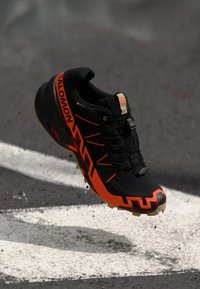 Chaussure de course noire et orange avec une tige texturée, un système de laçage proéminent et une semelle robuste. Logo visible sur le côté.