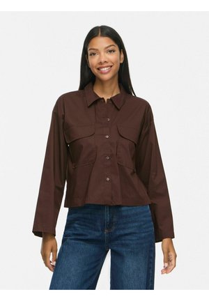 Femme aux longs cheveux noirs portant une chemise marron foncé à boutons avec des poches avant et un jean bleu, souriante sur un fond uni.