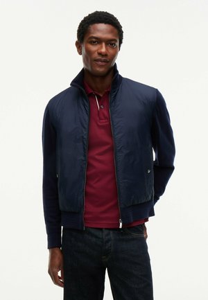 Mand iført en marineblå lynlåstrøje over en bordeaux pólotrøje og mørke jeans, stående med den ene hånd i lommen.