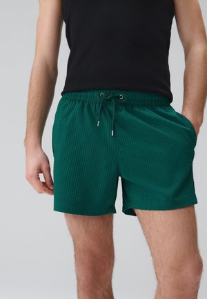 Mann trägt dunkelgrüne, gerippte Shorts mit Kordelzug und ein schwarzes ärmelloses Oberteil, steht mit einer Hand in der Tasche vor einfarbigem Hintergrund.
