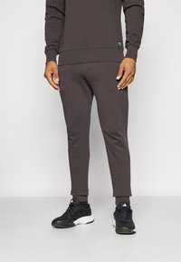Braune Baumwoll-Jogginghosen mit schmal zulaufenden Beinen und gerippten Bündchen, kombiniert mit schwarzen Sportschuhen. Einfaches Design, keine auffälligen Muster oder Logos.