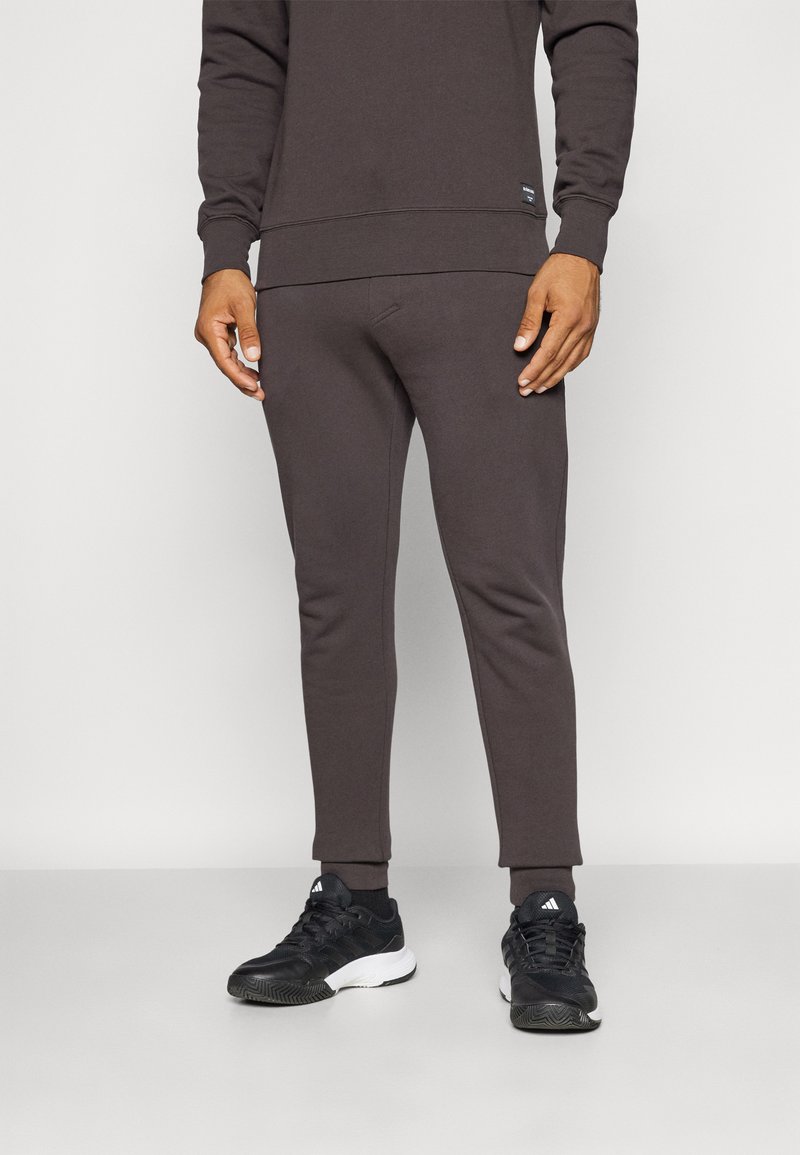 Braune Baumwoll-Jogginghosen mit schmal zulaufenden Beinen und gerippten Bündchen, kombiniert mit schwarzen Sportschuhen. Einfaches Design, keine auffälligen Muster oder Logos.