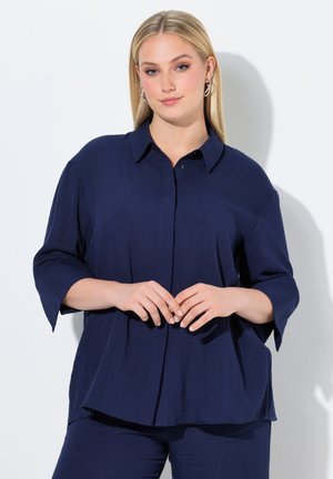 Blondynka o prostych włosach nosi luźną granatową koszulę zapinaną na guziki i pasujące spodnie, stoi na białym tle.