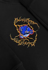 Zwarte sweatshirt met een geborduurd ontwerp van een blauw gezicht omringd door oranje bliksempatronen, wat een dynamisch visueel contrast creëert.