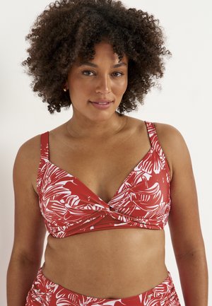 Woman with curly hair wearing a red and white tropical patterned bikini top, looking directly at the camera with a neutral expression.