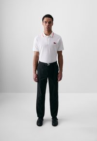 Polo blanc avec des détails de col rouge, arborant un petit logo. Assorti avec un pantalon noir ; le modèle se tient droit avec des chaussures noires.