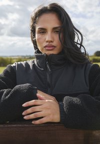 Schwarze Zip-Jacke mit einem kontrastierenden, flauschigen Material an den Ärmeln und dem Kragen. Die Hände des Models sind überkreuzt und zeigen natürliche Nägel.
