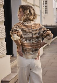 Pull en tricot avec un motif multicolore aux tons chauds. Présente un col montant, des poignets côtelés et une coupe décontractée. Associé à un pantalon de couleur claire.