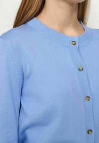 Ljusblå stickad cardigan med rund hals, med en full knappslå av bruna knappar och ribbade detaljer längs kanterna.