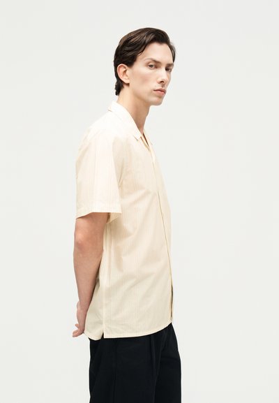 Νέος άνδρας όρθιος πλαγίως με τα χέρια πίσω από την πλάτη, φορώντας μπεζ πουκάμισο με κοντά μανίκια και μαύρο παντελόνι, ουδέτερο φόντο.