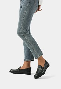 Mocassini in pelle nera con un accento in catena metallica, suola piatta e punta rotonda, abbinati a jeans azzurri chiari, leggermente cropped.