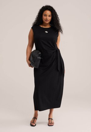 Žena kučeravých vlasov v bezrukávových čiernych midi šatách s viazačkou na boku, v čiernych sandáloch, so strieborným náhrdelníkom a drží čiernu listovú kabelku.