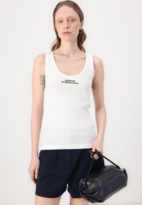 Valge sansvarrukatega T-särk, millel on "BARBOUR INTERNATIONAL" logo, koos tumesiniste lühikeste pükstega ja musta ning tumesinise käekotiga. Sile tekstuur.