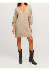 Beige neulemekko, jossa on V-aukko, pitkät pallotakkihihat ja ribattu helma; yhdistetty mustiin cowboy-saappaisiin, joissa on kohokuvioitu yksityiskohta.