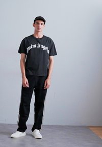 Melnas kokvilnas T-kreklu ar "Palm Angels" logo baltā krāsā, apvienots ar melniem biksēm, kurām ir baltas sānu svītras, un baltām sporta kurpēm. Modelis stāv taisni.