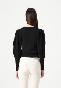 Maglione nero con maniche in pizzo strutturato e gonfio, fondo a costine aderente e scollatura scalloppata. Indossato con jeans di colore chiaro.