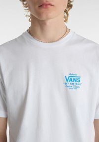 Vit bomull t-shirt med en blå grafisk logotyp på vänster bröstområde, rund halsringning och korta ärmar.