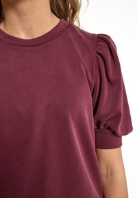 Mujer que lleva una blusa corta de manga con volantes en color burdeos y cuello redondo, mostrada desde el cuello hasta la parte superior del brazo.
