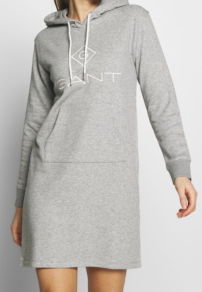 Femme portant une robe sweat à capuche gris clair avec poche avant et logo GANT sur la poitrine sur un fond uni.