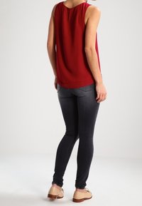 Femme se tient de dos, portant un blouse sans manches rouge, un jean skinny noir et des chaussures oxford beiges sur un fond clair et uni.