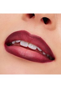 Dyp bærfarget matt leppestift på fyldige lepper, som viser den jevne teksturen og veldefinerte formen. Hvite tenner er svakt synlige.