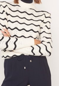 Pull en tricot blanc avec des rayures ondulées noires et un col montant. Poignets et ourlet côtelés, mettant en valeur un motif ajouré et un tissu texturé.