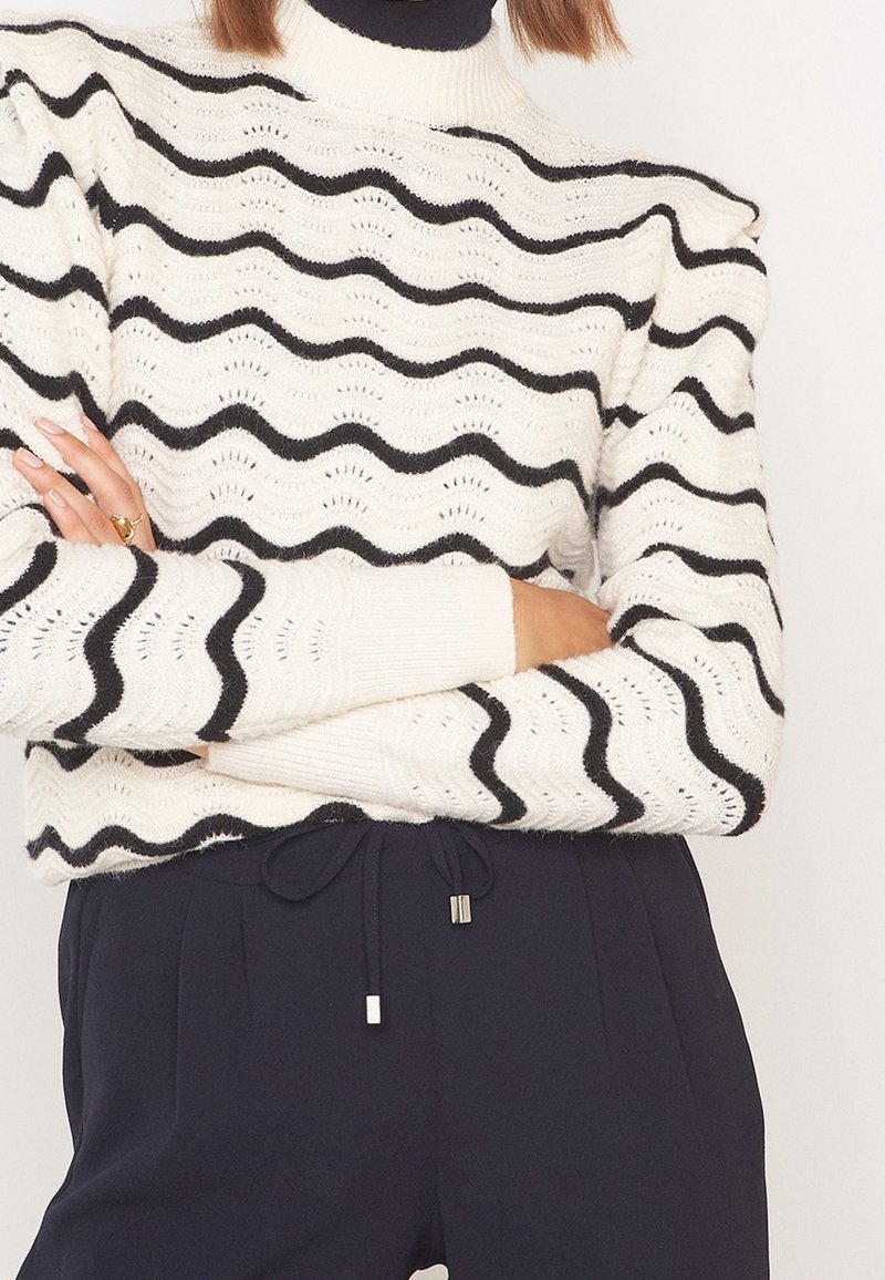 Pull en tricot blanc avec des rayures ondulées noires et un col montant. Poignets et ourlet côtelés, mettant en valeur un motif ajouré et un tissu texturé.