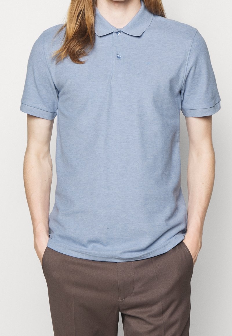 Personne portant un polo bleu clair à manches courtes rentré dans un pantalon marron, les mains dans les poches, sur un fond uni.