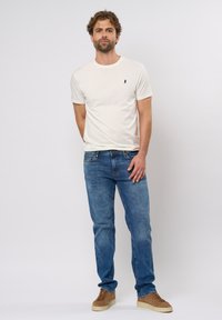 Beež crew-neck t-särk väikese tumesinise logoga, sinised denimist teksad ja helepruunist nahast tossud; lõdva lõikega ja sujuvad tekstuurid kogu ulatuses.