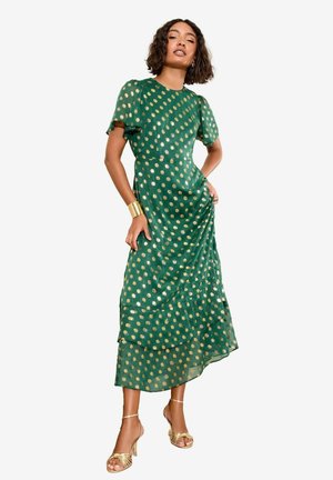 Grünes Kleid mit kurzen Ärmeln, das mit goldenen Punkten verziert ist, Schichten-Design und fließender Textur. Trägt goldene Riemchen-Heels.