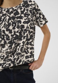 Inimene, kes kannab must-valge abstraktse mustriga lühikeste varrukatega särki ja tumedaid täisvildis teksaseid, reguleerib musta vööd neutraalsel taustal.