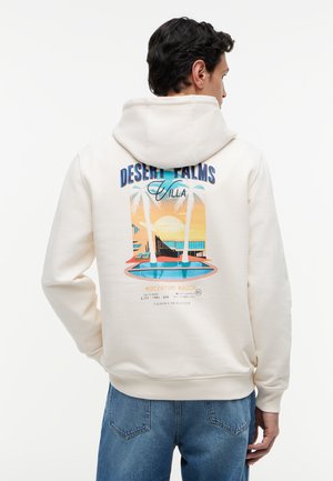 Férfi világos kapucnis pulóverben, amelynek hátán színes naplemente, pálmafák és egy medence ábrája látható, felirat: "Desert Palms Villa", kék farmernadrágot visel.