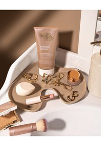 Lotion autobronzante progressive Bondi Sands avec des outils de maquillage comprenant un rouge à lèvres, une éponge pour le visage, un recourbe-cils, de la poudre, un pinceau, des bagues et des boucles d'oreilles sur un plateau de salle de bain.
