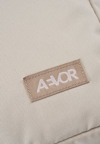 Beige Textiltaschenoberfläche mit strukturierter Oberfläche und einem gestickten, rechteckigen, beigen Etikett mit dem Markennamen "A=VOR" in weißer Stickerei.