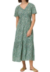 Femme portant une robe midi verte à fleurs, col en V, manches courtes et des sandales beiges à enfiler, debout avec une main posée sur la robe.