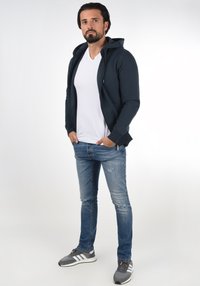 Navyblauer Kapuzenzipper über einem weißen V-Ausschnitt-Shirt, kombiniert mit hellblauen, ausgewaschenen Jeans und grauen Sneakers mit weißen Akzenten.