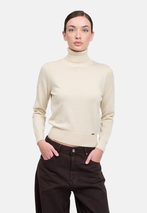 Donna con capelli scuri che indossa un maglione crema a collo alto e pantaloni larghi scuri, in piedi con le mani nelle tasche su uno sfondo bianco semplice.